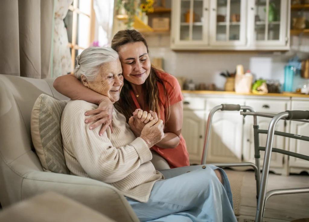 Woman sitting on a couch with a walker beside her, embracing a caregiver who is standing on the floor
