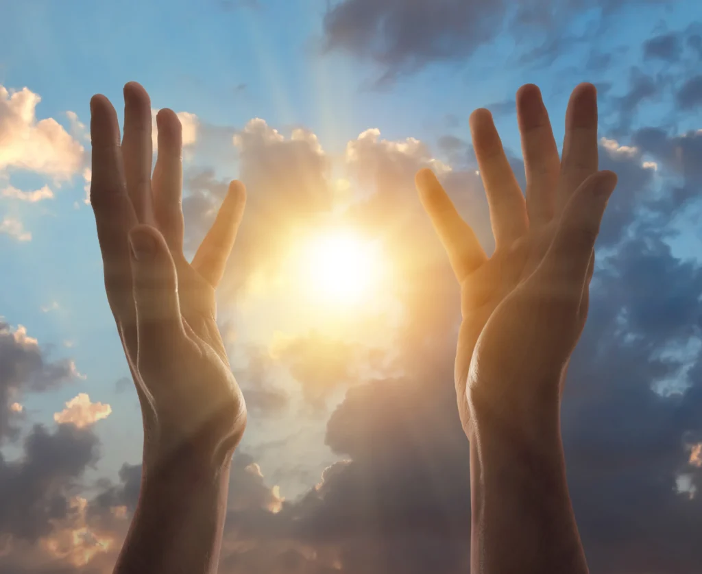 Two hands reaching up to the sky with the sun shining through the clouds, symbolizing the power of prayer