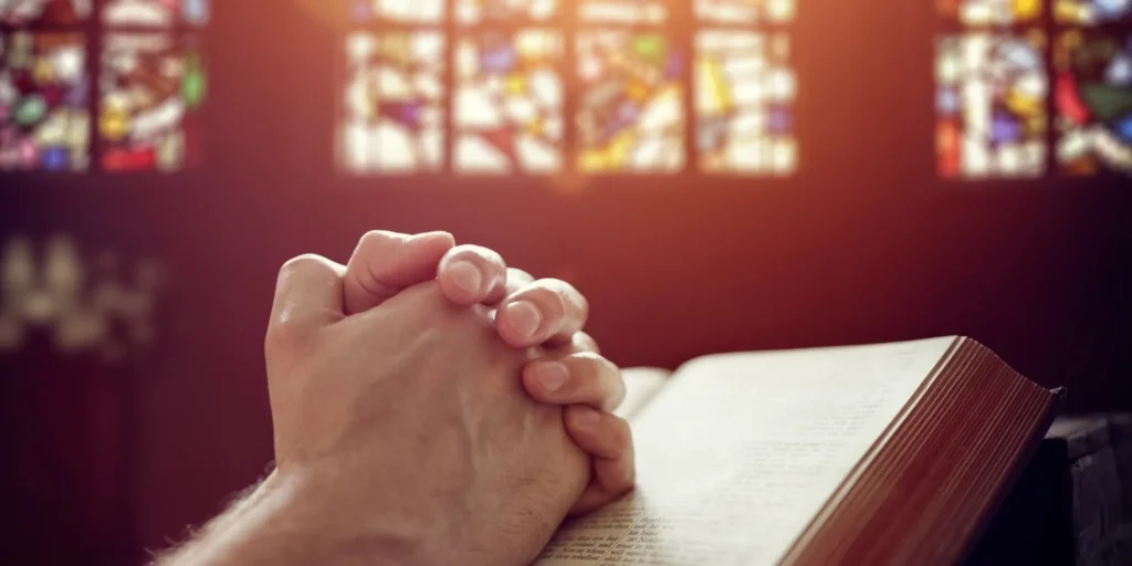 Person with their hands clasped in prayer, reading a book in front of a stained glass window