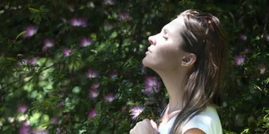 Woman standing in front of a bush with her eyes closed, surrounded by trees and flowers