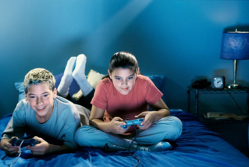 Two children sitting on a bed, each holding a joystick and playing video games