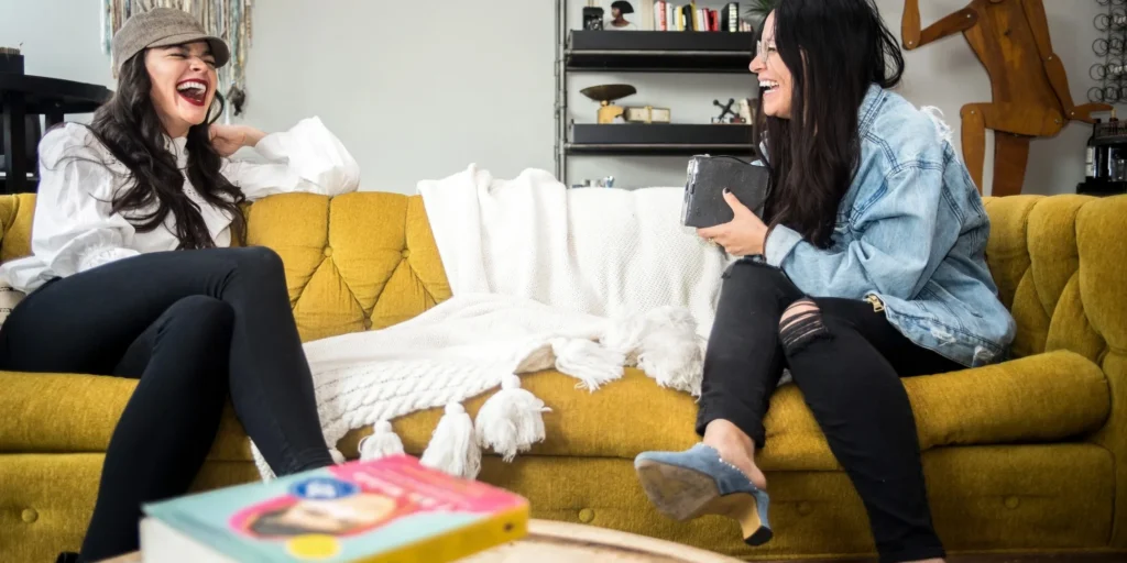 Two women sitting on a yellow couch, laughing and talking to each other
