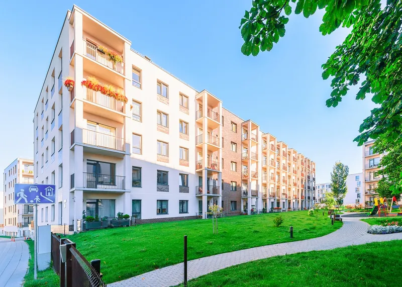 Large apartment building with a green lawn in front of it, surrounded by trees, plants, flowers, and a fence