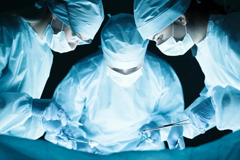 Three surgeons wearing masks, caps, gloves, and coats, performing surgery in an operating room
