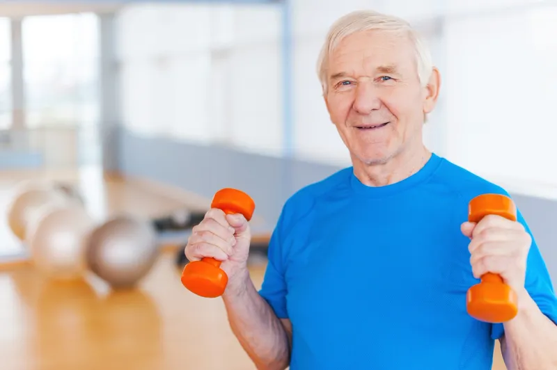 Older man wearing a t-shirt, holding two orange dumbbells in his hands, with a blurred background