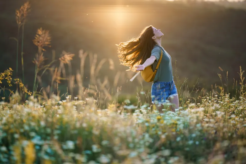 Woman standing in a field of flowers with her arms outstretched, wearing a bag
