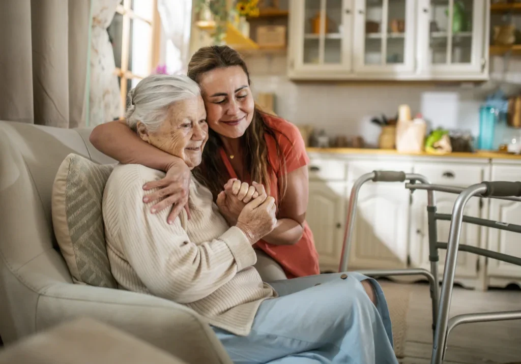 Woman sitting on a couch with a walker beside her, embracing a caregiver who is standing on the floor