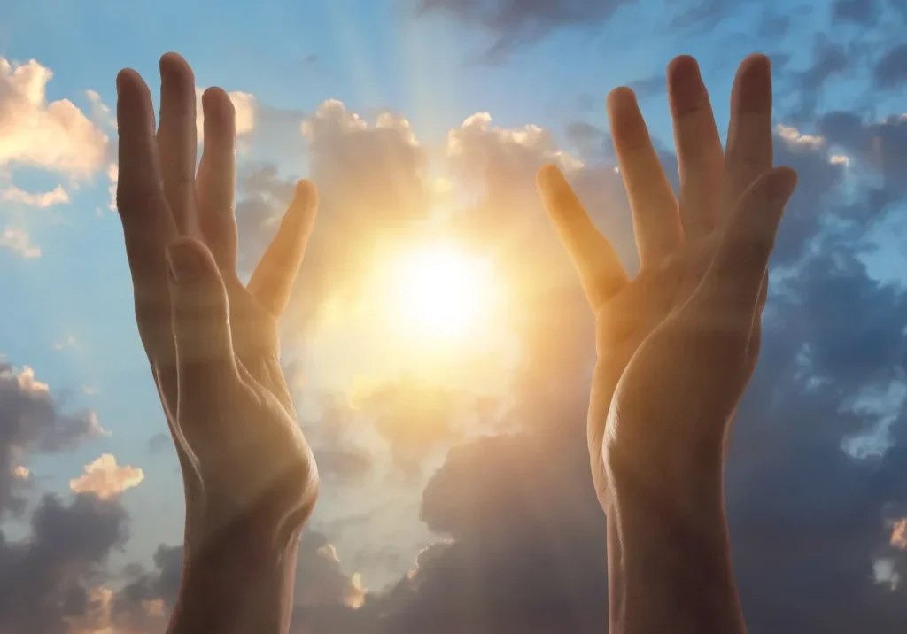 Two hands reaching up to the sky with the sun shining through the clouds, symbolizing the power of prayer