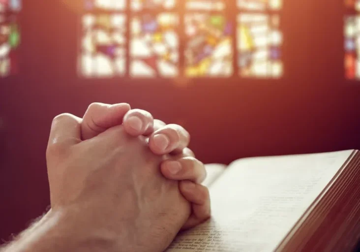 Person with their hands clasped in prayer, reading a book in front of a stained glass window
