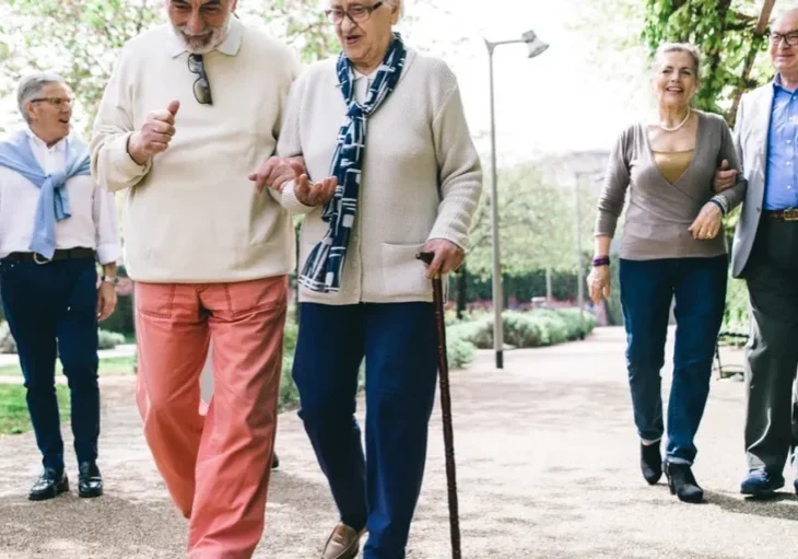 Group of older people walking down a path in a park, surrounded by trees, plants, and grass