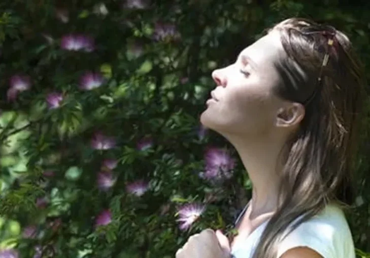 Woman standing in front of a bush with her eyes closed, surrounded by trees and flowers