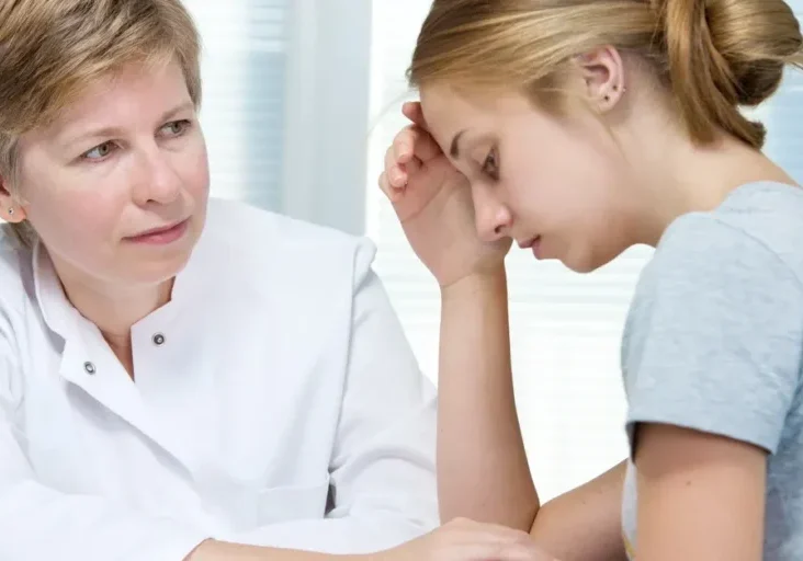 Woman sitting at a table with her head in her hands, while a doctor stands beside her