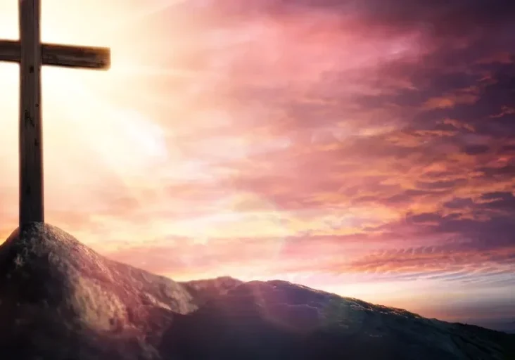 Cross atop a mountain with the sun setting in the background, surrounded by clouds in the sky