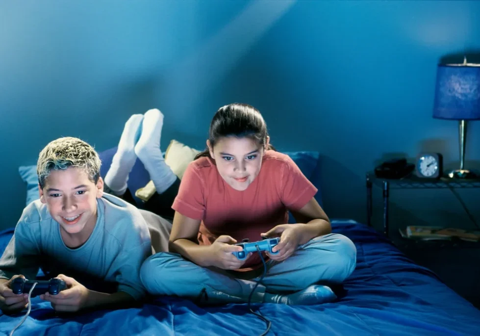 Two children sitting on a bed, each holding a joystick and playing video games
