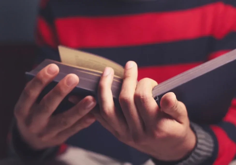 Person wearing a red and t-shirt, holding a book in their hands and reading it intently