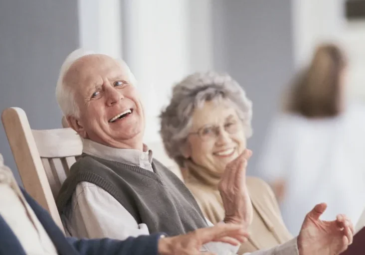Older couple sitting in chairs, and laughing, with a woman standing in the background