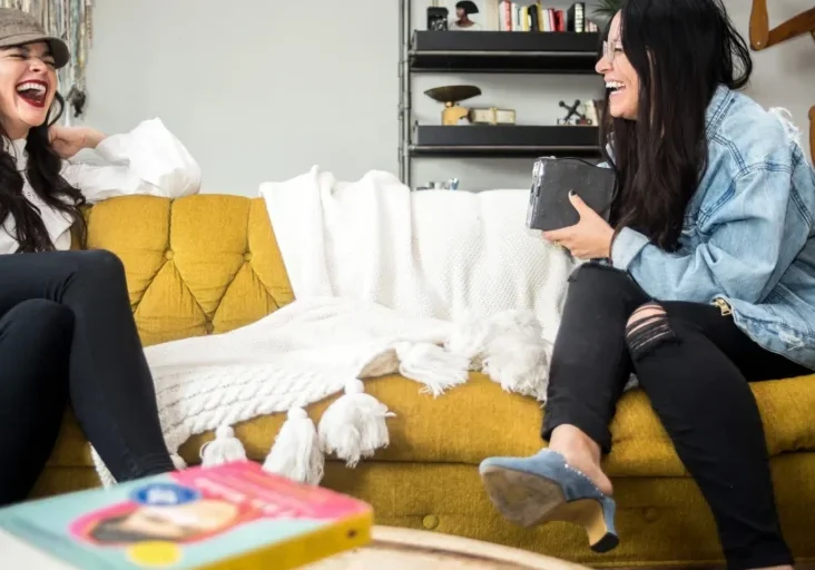 Two women sitting on a yellow couch, laughing and talking to each other
