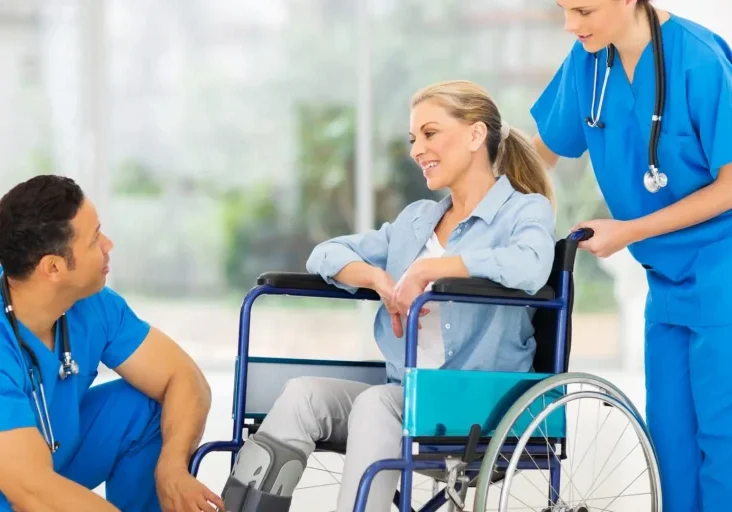 Woman in a wheelchair being assisted by two nurses wearing stethoscopes, with a blurred background