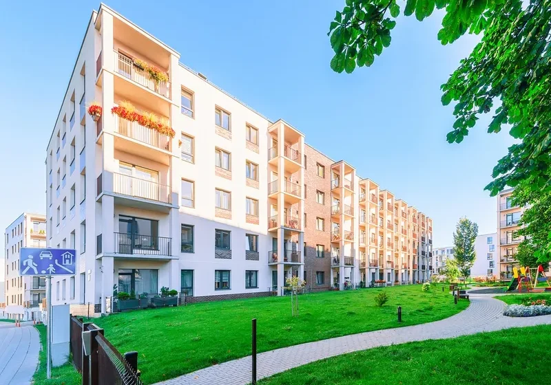Large apartment building with a green lawn in front of it, surrounded by trees, plants, flowers, and a fence