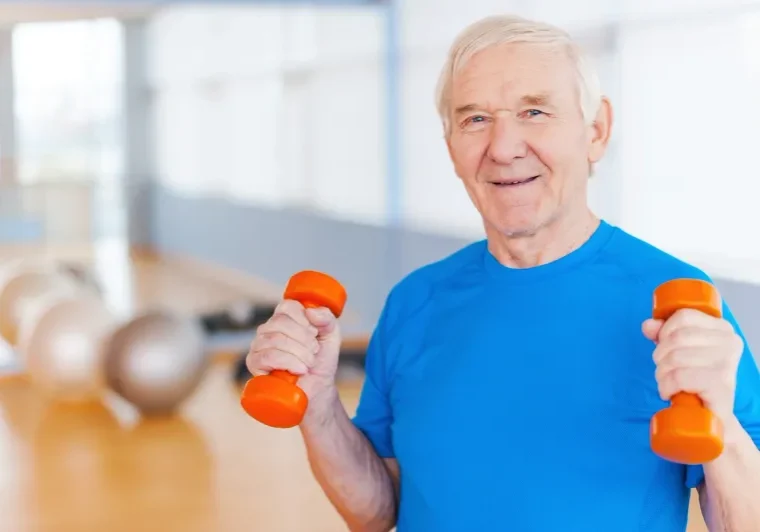 Older man wearing a t-shirt, holding two orange dumbbells in his hands, with a blurred background