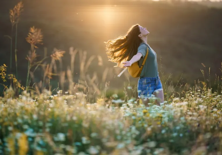 Woman standing in a field of flowers with her arms outstretched, wearing a bag